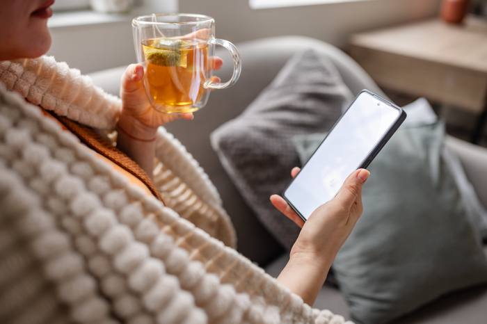 A person under a blanket holding a mug of tea, phone resting nearby.